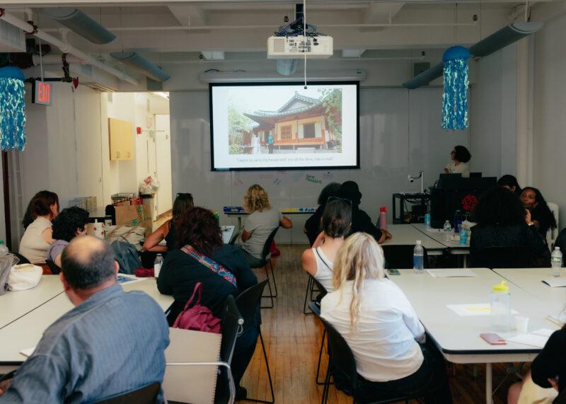 A group of people sit at tables in a classroom, watching a presentation displayed on a screen.