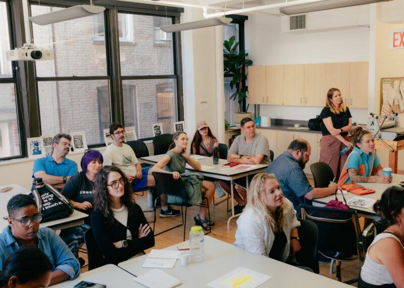 A group of adults sit at desks in a bright classroom. Some take notes, others look ahead attentively.