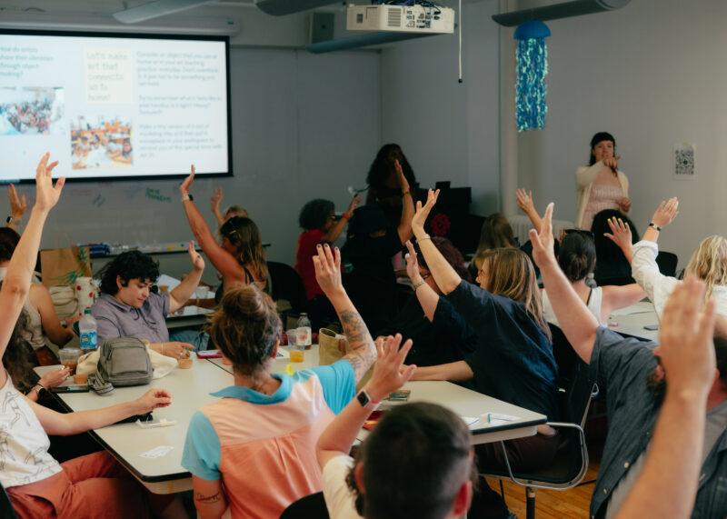 People seated around tables in a classroom, raising their hands while a presenter stands in front near a screen displaying a presentation.