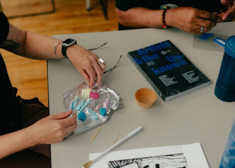 Two people sit at a table with modeling clay, wooden sticks, a cupcake liner, a book titled "Artists & the Unknown," and a drawing in black ink on white paper.