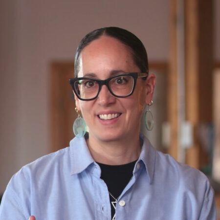 Dyani White Hawk with dark hair pulled back, wearing large glasses, circular earrings, and a light blue collared shirt, smiles at the camera indoors.