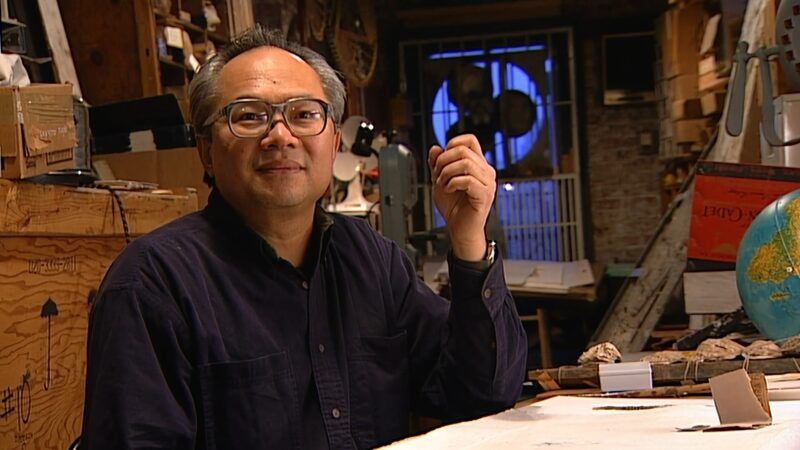 Mel Chin wearing glasses and a dark shirt sits in a cluttered workshop, surrounded by various tools and objects.